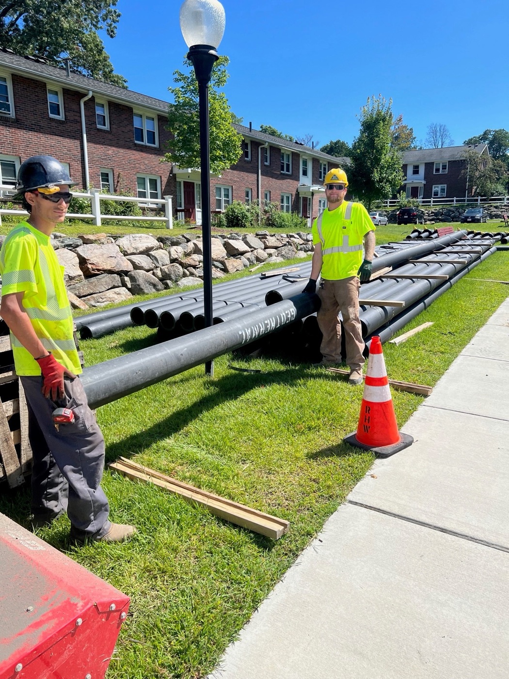 Two construction workers in safety gear next to rows of black HDPE ambient-loop pipe in front of a brick apartment complex.